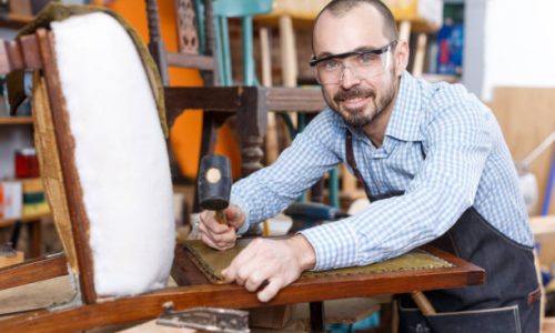 Positive craftsman engaged in repair of vintage furniture, reupholstering chair in workshop